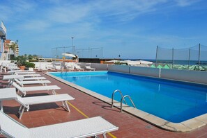 Una piscina en la azotea con sillones blancos, cielo azul despejado y vista al mar.