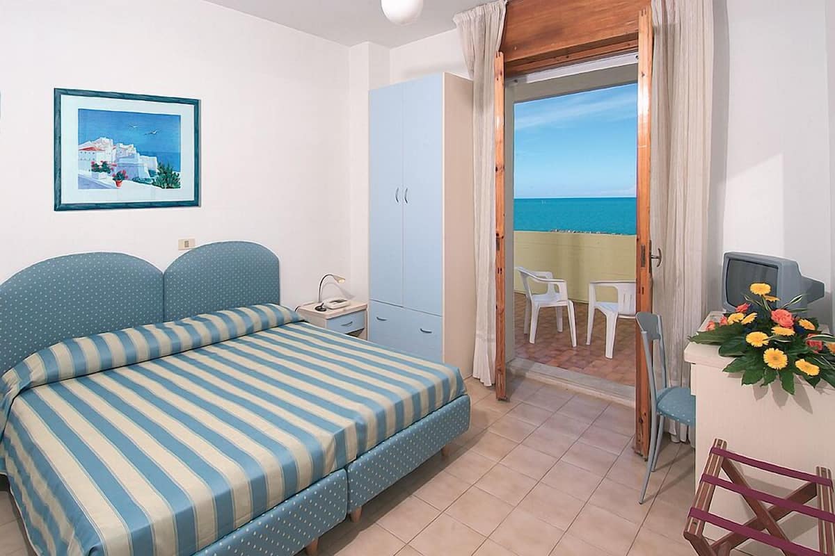 A bedroom with a bed, a wardrobe, a desk with a chair, and a balcony with a view of the sea.