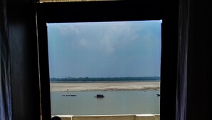 Vista desde un balcón sobre un río con barcos, una playa de arena y un horizonte urbano lejano.