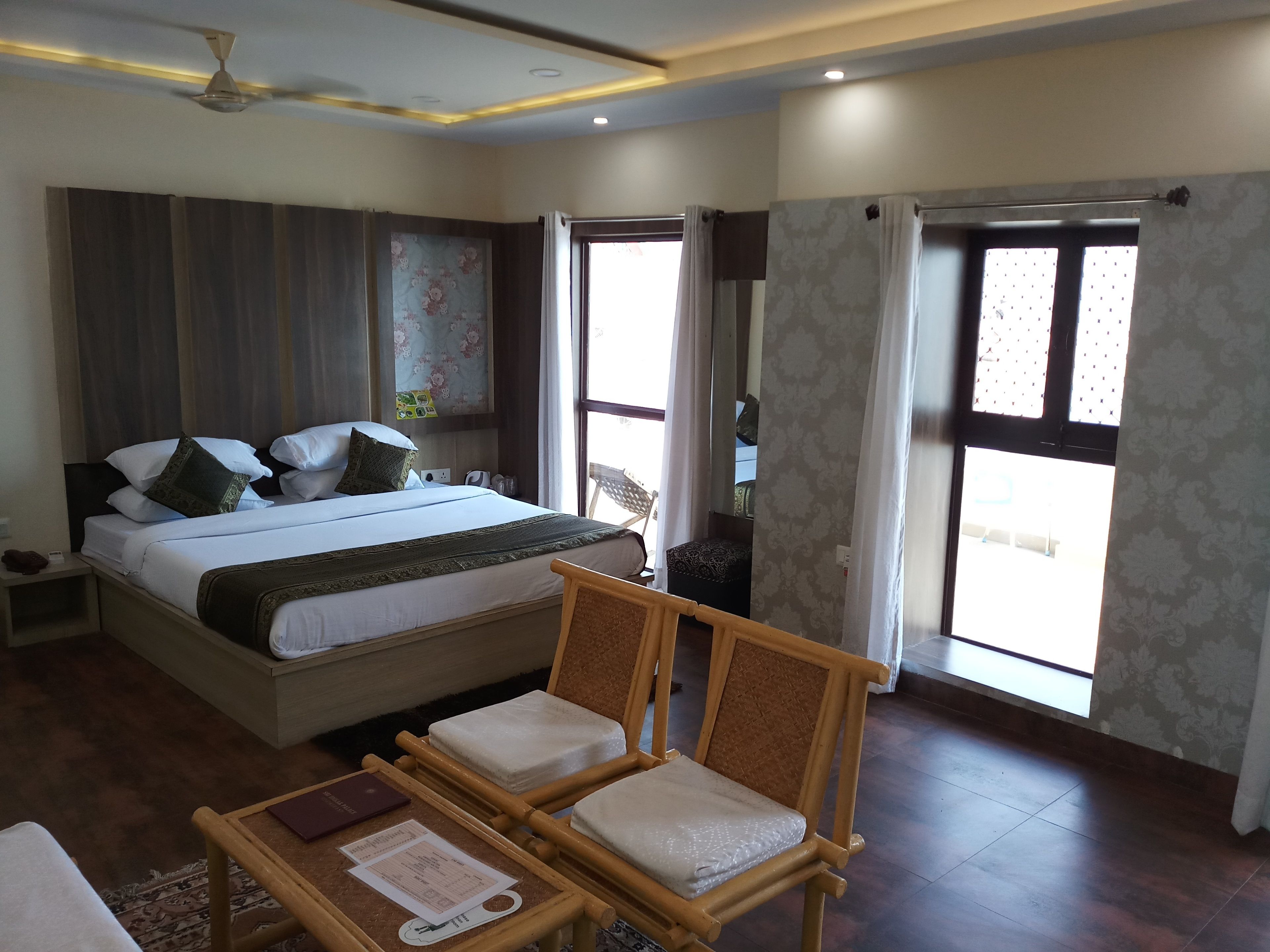 A hotel room with a large bed, a wooden chair, a small table with a booklet, and a ceiling fan.