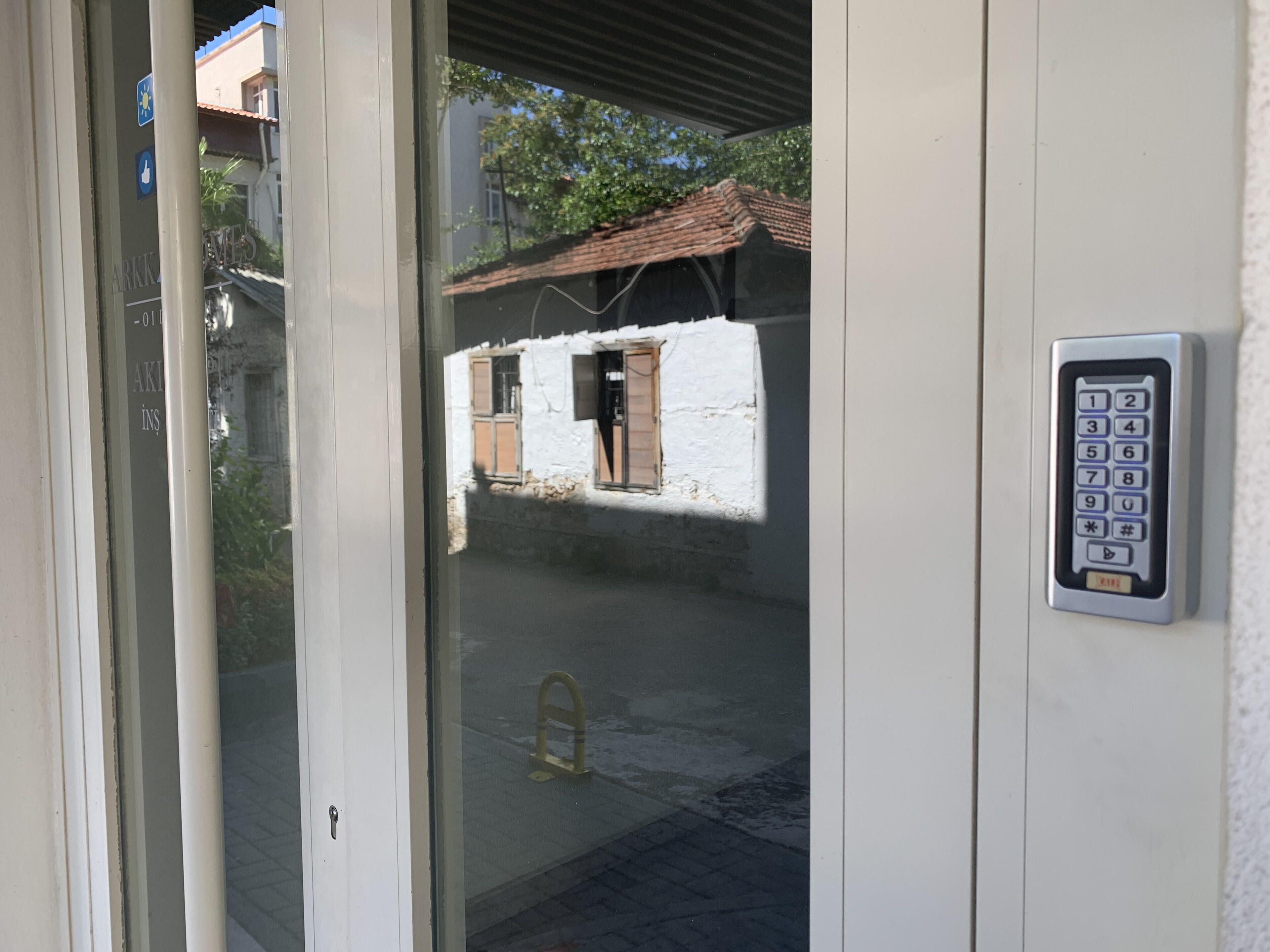 Una cerradura electrónica con teclado, una puerta de cristal y una vista de un edificio con tejado de azulejos y una ventana con contraventanas de madera.