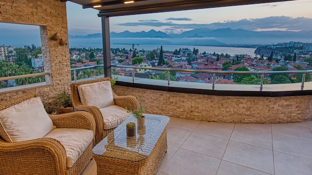 A balcony with wicker furniture, a glass table, and a view of a city and mountains. - Arkk Homes (Antalya)