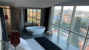 A modern bedroom with a large window, a bathtub, and a view of the city. - Arkk Homes (Antalya)