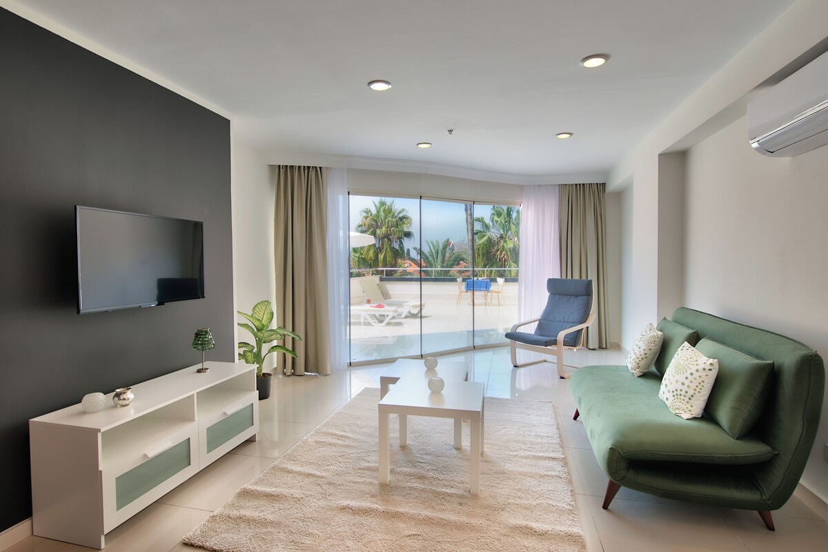 A modern living room with a flat-screen TV, a green sofa, a white coffee table, and a large sliding glass door leading to a balcony with outdoor furniture.