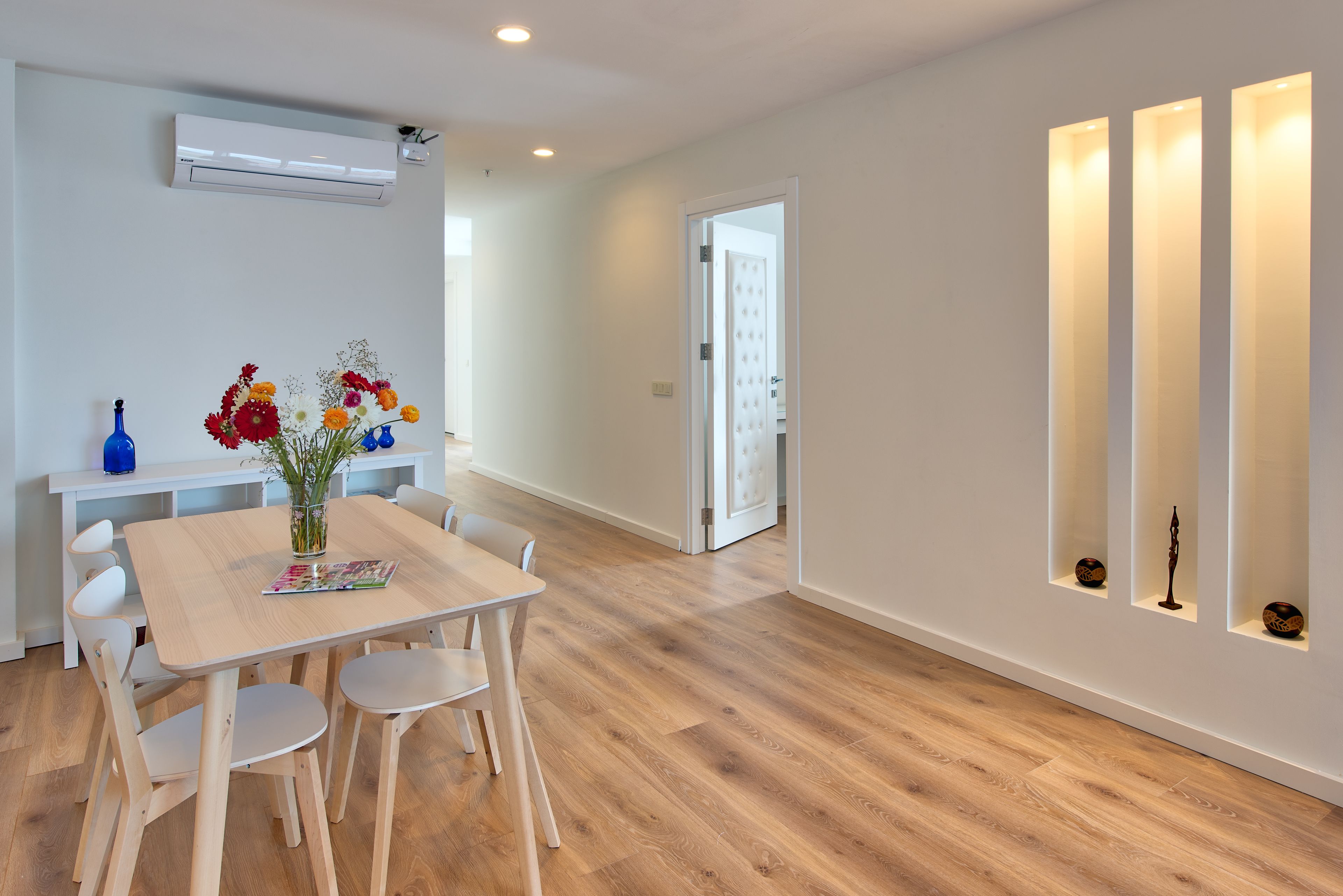 A modern interior with a wooden floor, a dining table with chairs, a vase of flowers, and a white wall with shelves.