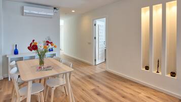 A modern interior with a wooden floor, a dining table with chairs, a vase of flowers, and a white wall with shelves.