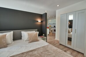 A modern bedroom with a large bed, a wardrobe, and a kitchenette in the background. - Arkk Homes (Antalya)