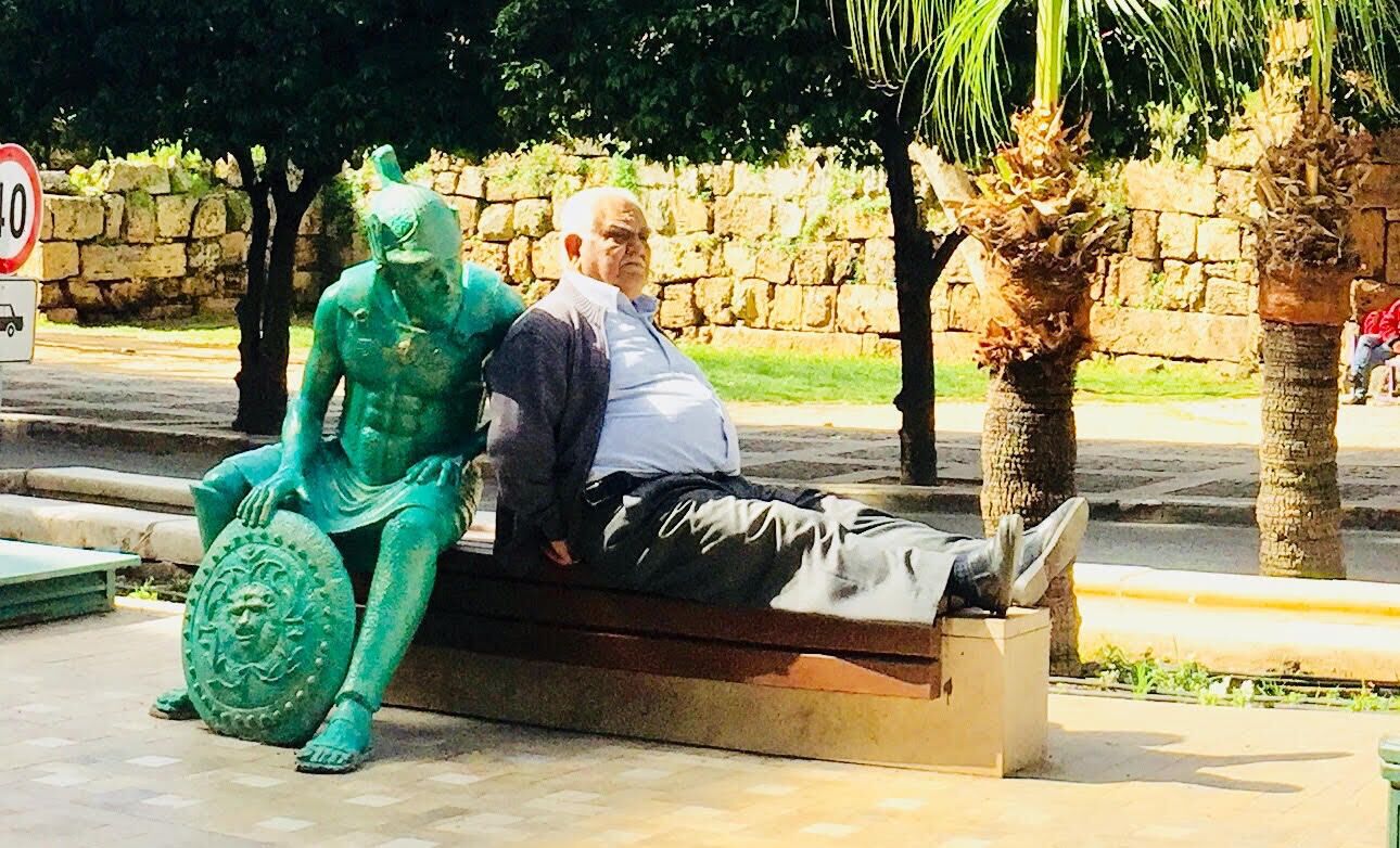 Une statue représentant une figure assise, tenant un bouclier, avec un banc et un mur de pierre en arrière-plan.
