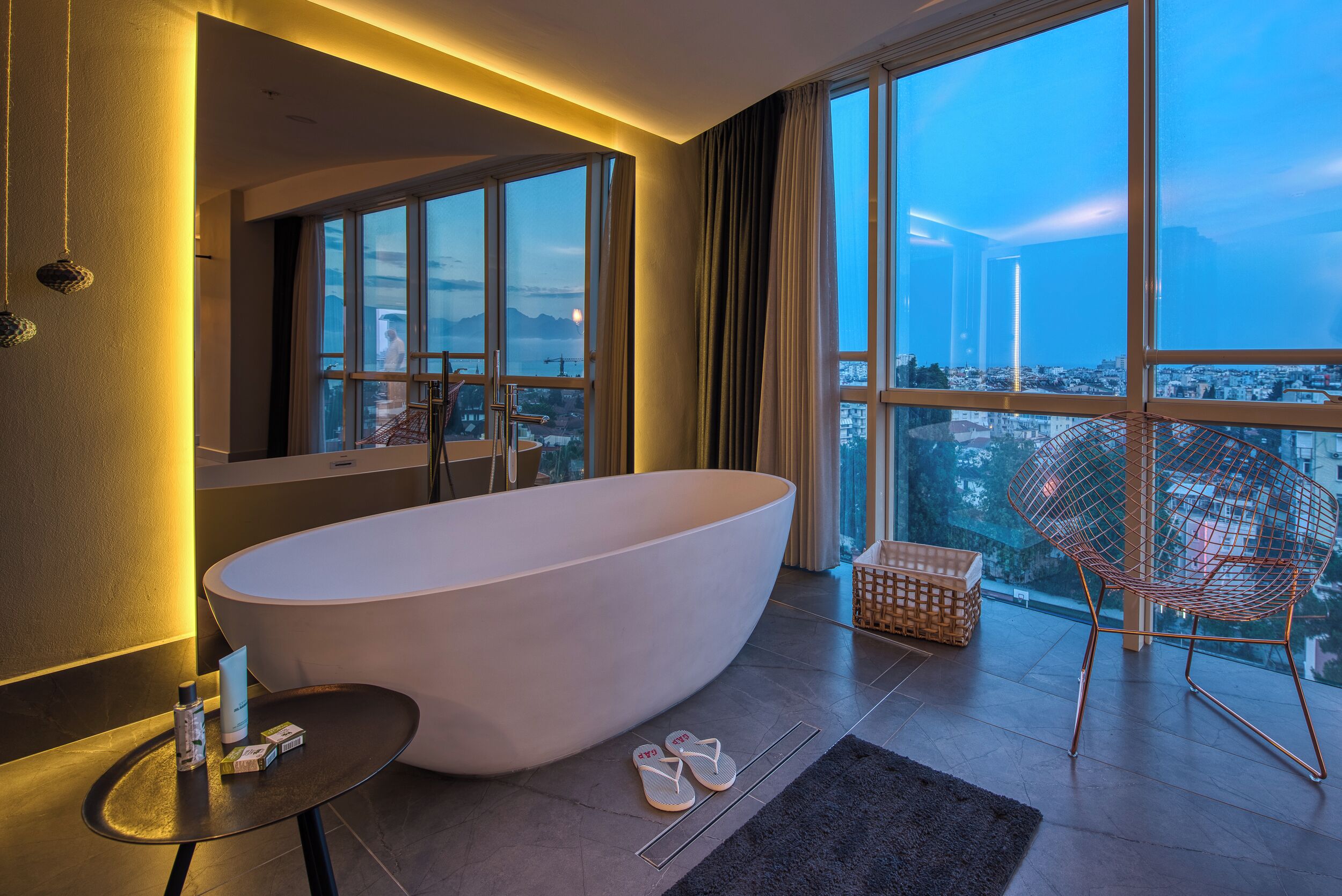Un baño moderno con una bañera independiente, un espejo grande y vistas a un paisaje urbano a través de amplios ventanales.