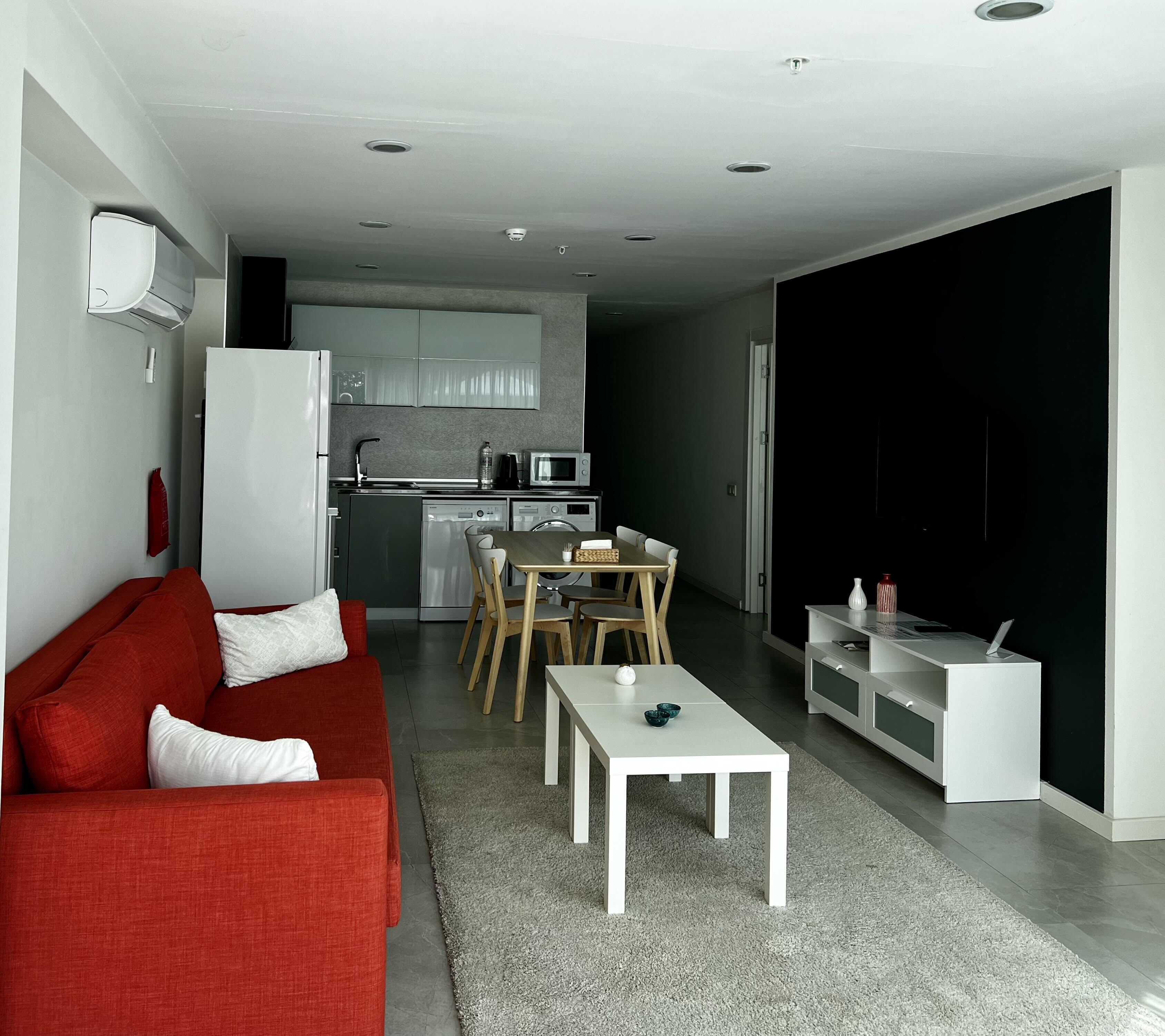 Een moderne woonkamer met een rode bank, een witte salontafel en een eethoek met een tafel en stoelen. In de achtergrond is een keuken zichtbaar met een koelkast, magnetron en spoelbak.