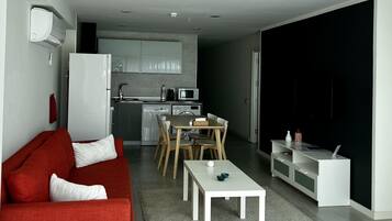 Een moderne woonkamer met een rode bank, een witte salontafel en een eethoek met een tafel en stoelen. In de achtergrond is een keuken zichtbaar met een koelkast, magnetron en spoelbak.