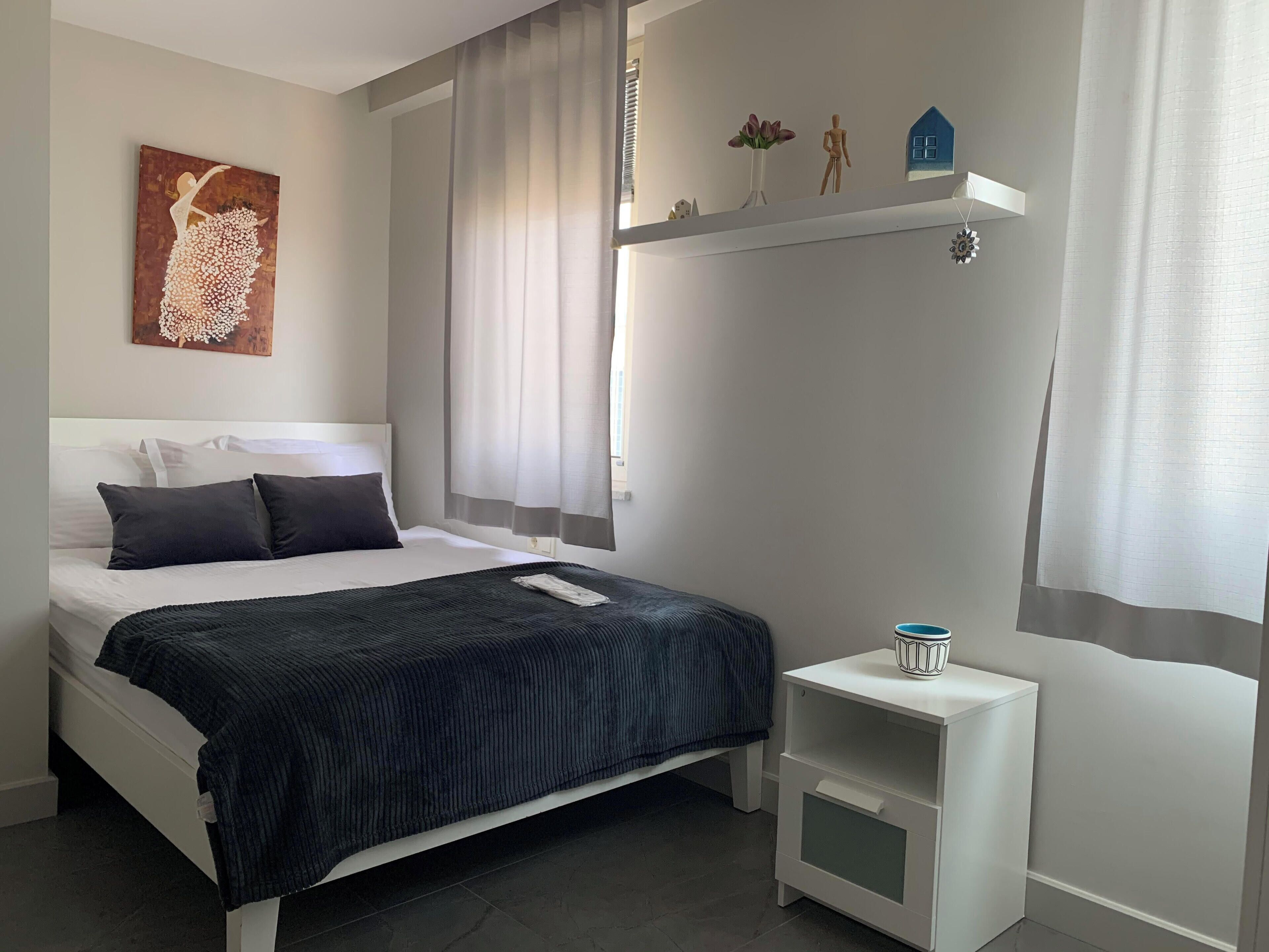 Une chambre à coucher bien rangée, avec un lit, une table de chevet, une étagère ornée d’objets décoratifs et une fenêtre avec des rideaux.