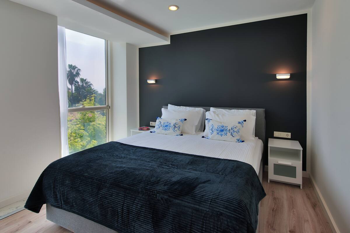 A modern bedroom with a large bed, two bedside tables, and a view of greenery outside.