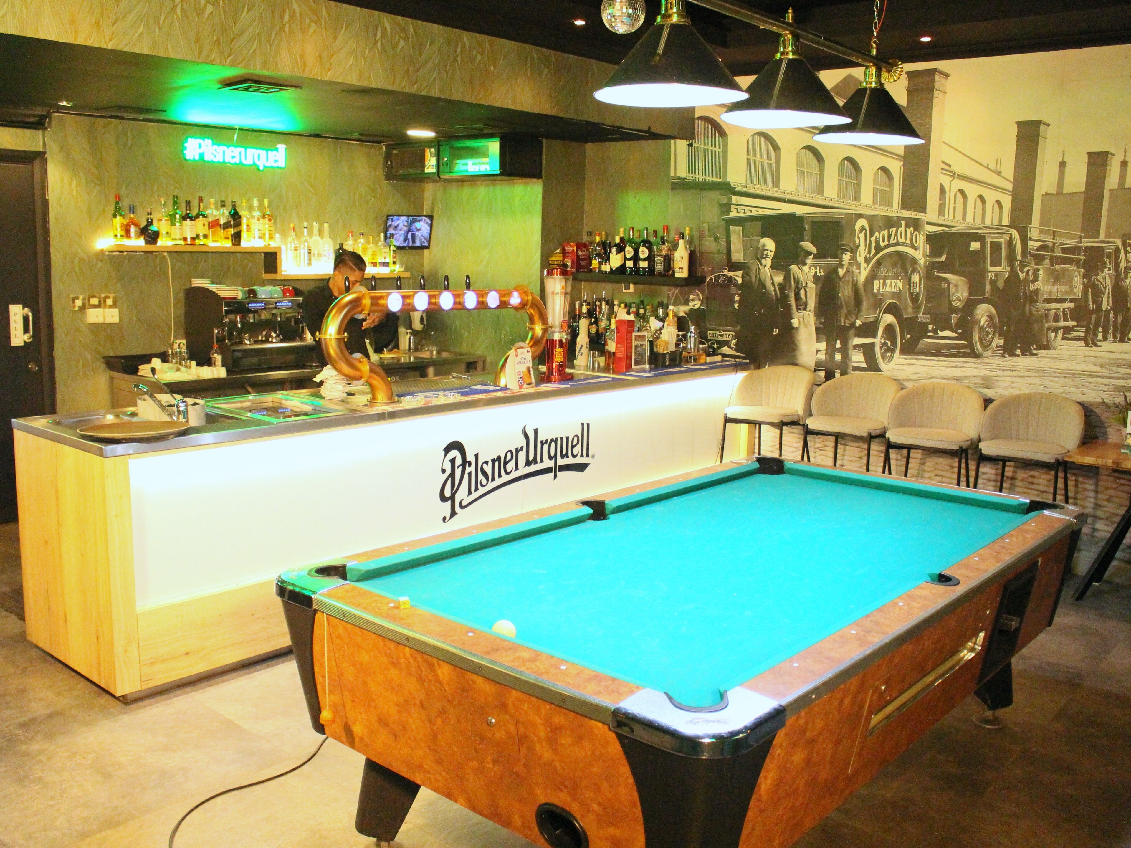 A billiards table in a bar area with a Pilsner Urquell sign, a bar counter with various bottles, and a vintage car mural in the background.