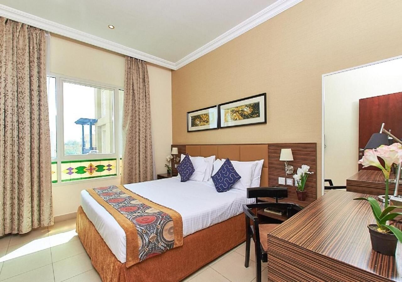 A hotel room with a bed, a desk, a chair, a window with curtains, and a wall with framed pictures.
