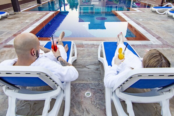 Dos personas descansando en sillones de piscina junto a una pileta con piso de baldosas y agua cristalina.