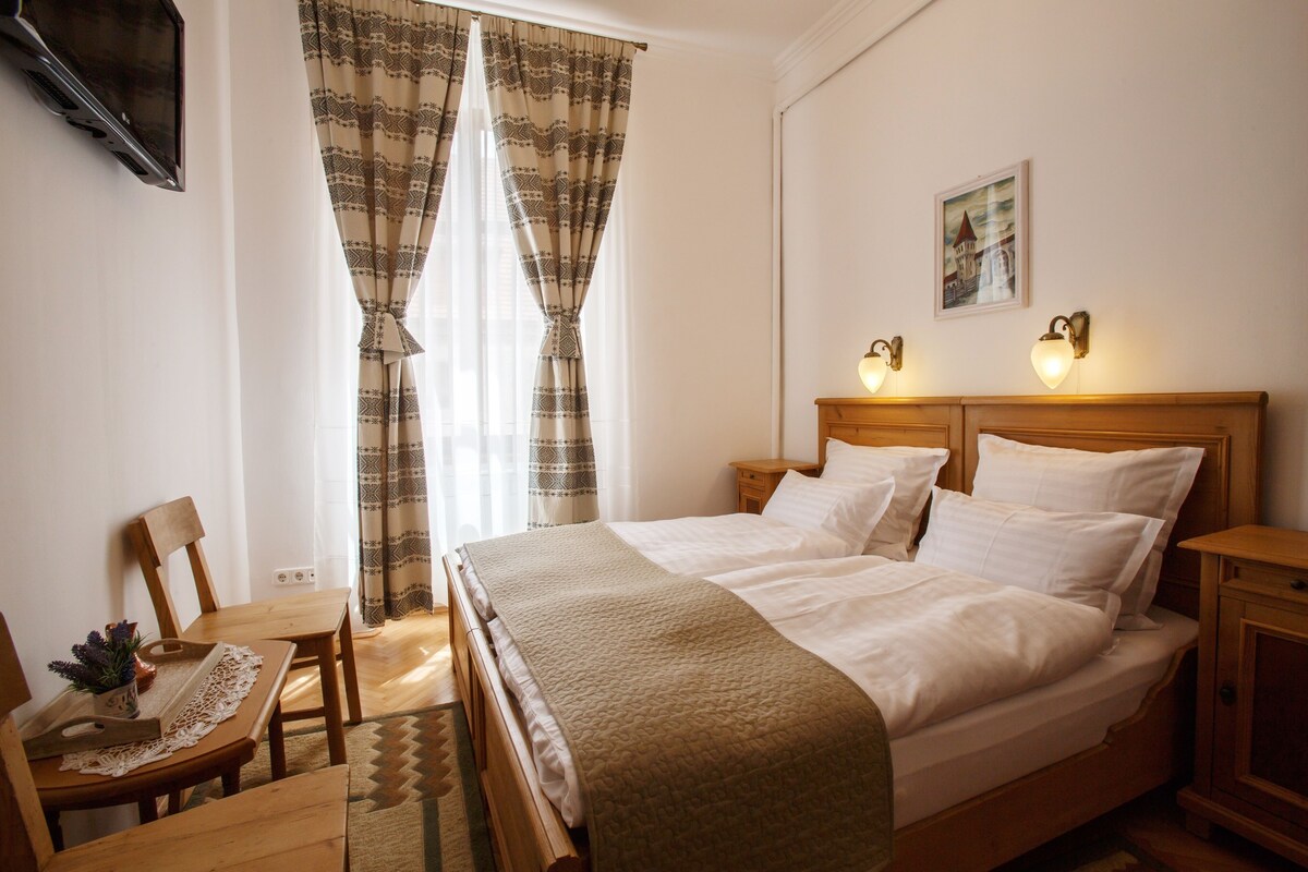 Una habitación de hotel con una cama, dos mesitas de noche, un televisor montado en la pared, una ventana con cortinas estampadas, una silla de madera y una pequeña mesa con una bandeja.