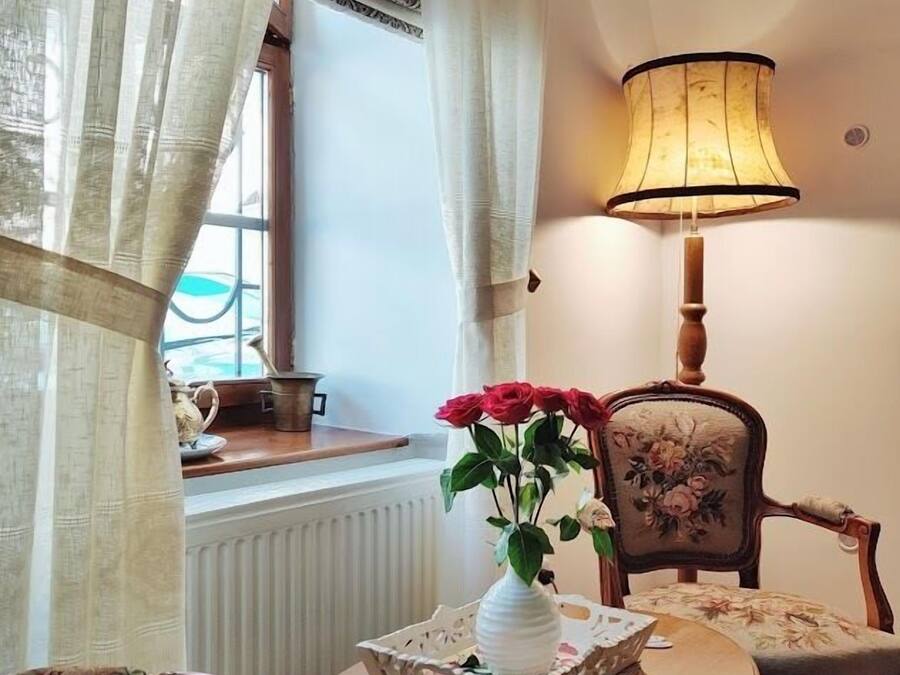 A cozy room with a round wooden table, a white vase with red flowers, a lamp, and a chair with floral upholstery.