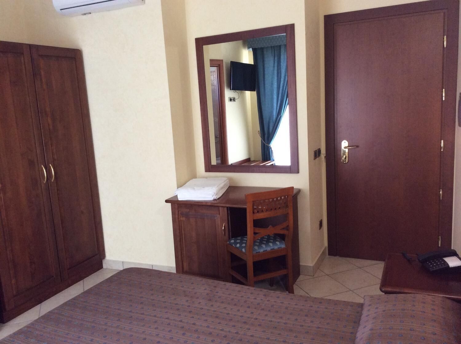 Een slaapkamer met een bed, een houten garderobe, een klein bureau met stoel, een spiegel en een deur.