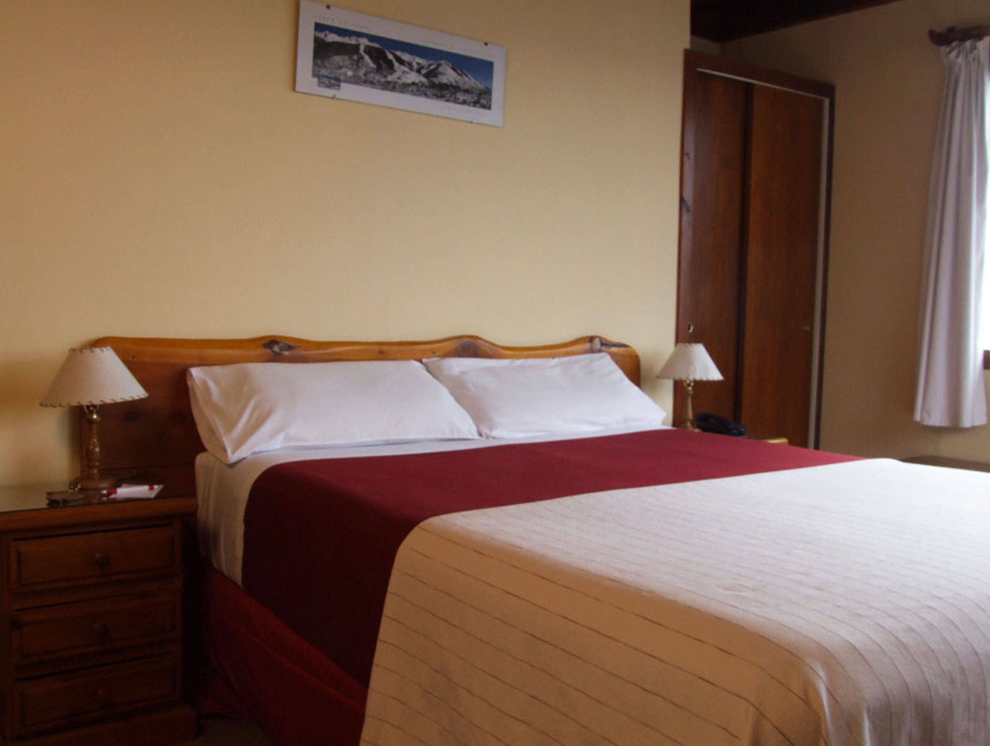 Una habitación de hotel con cabecera de madera, una cama grande con manta roja, dos lámparas de noche, una mesita de noche de madera y una puerta a la derecha.