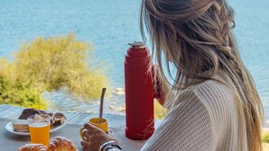 A person sitting at a table with breakfast items, a red thermos, and a view of a lake and mountains. - Hosteria Ventus Patagonia (San Carlos de Bariloche)