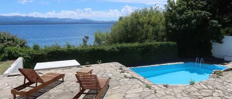 A pool with a view of the sea, surrounded by stone tiles and wooden loungers.