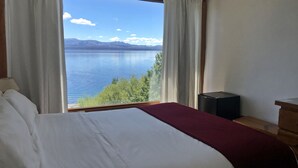 A hotel room with a large bed, a view of a lake, and a speaker on a wooden table. - Hosteria Ventus Patagonia (San Carlos de Bariloche)