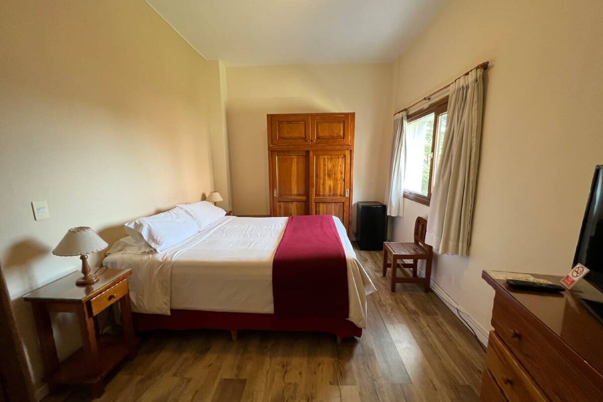 Ein Schlafzimmer mit Bett, Holzschrank, Nachttisch mit Lampe, Stuhl und Fernseher auf einer Kommode.