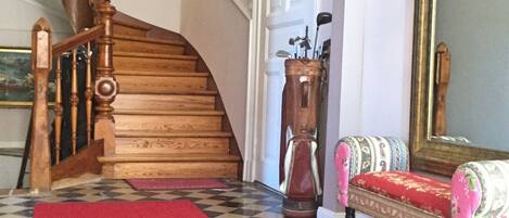 A staircase with wooden railings, a red carpet, a checkered floor, a bench with floral upholstery, and a wall-mounted mirror.