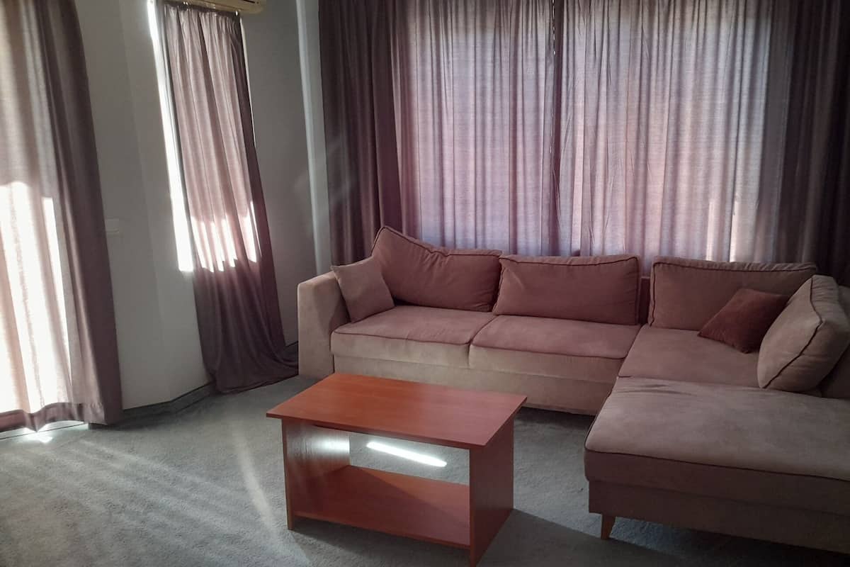 Una sala de estar con un sofá seccional color beige, una mesa de centro de madera y vistas a un edificio a través de cortinas traslúcidas.
