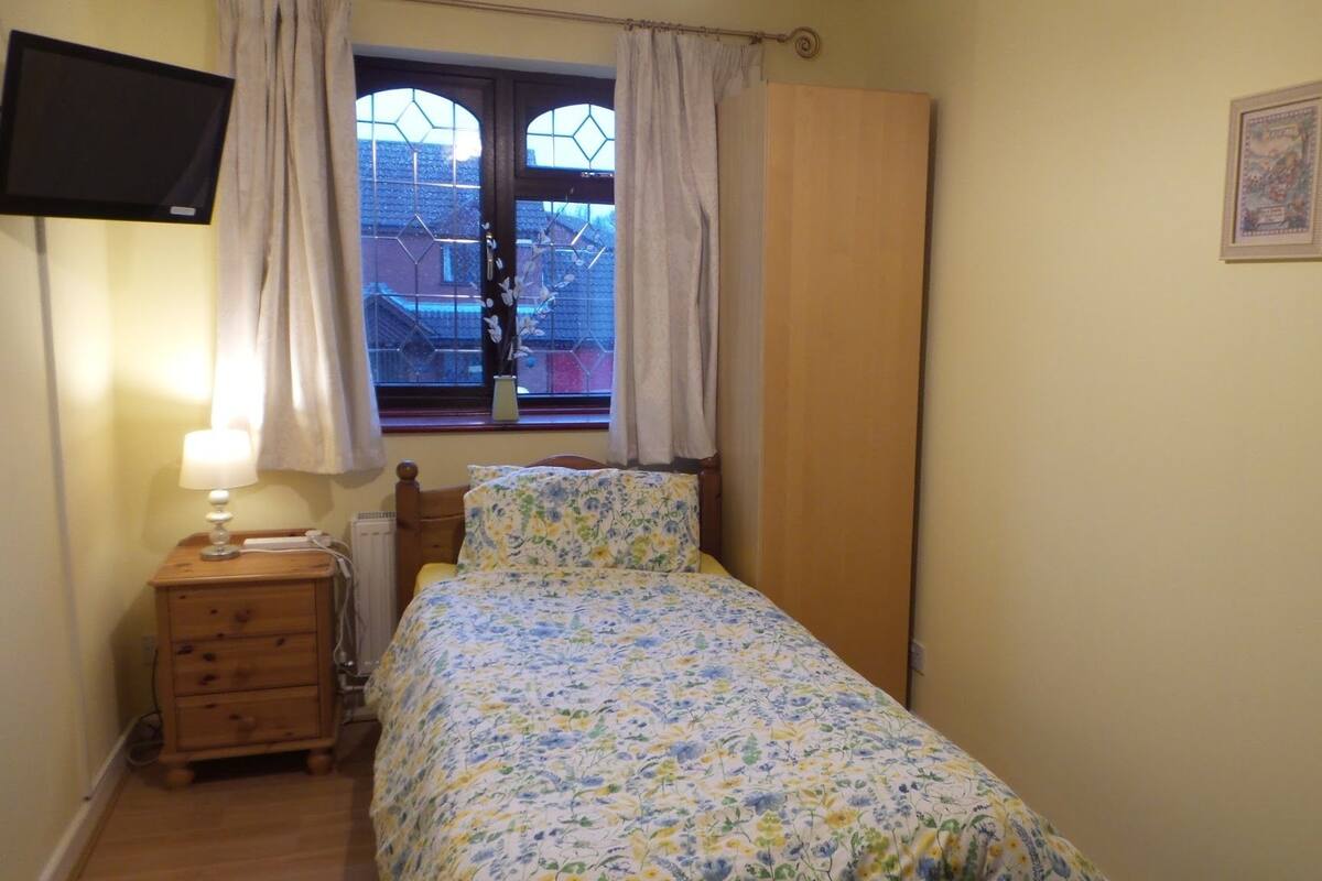 A bedroom with a bed, a nightstand, a lamp, a window with curtains, a radiator, and a framed picture on the wall.