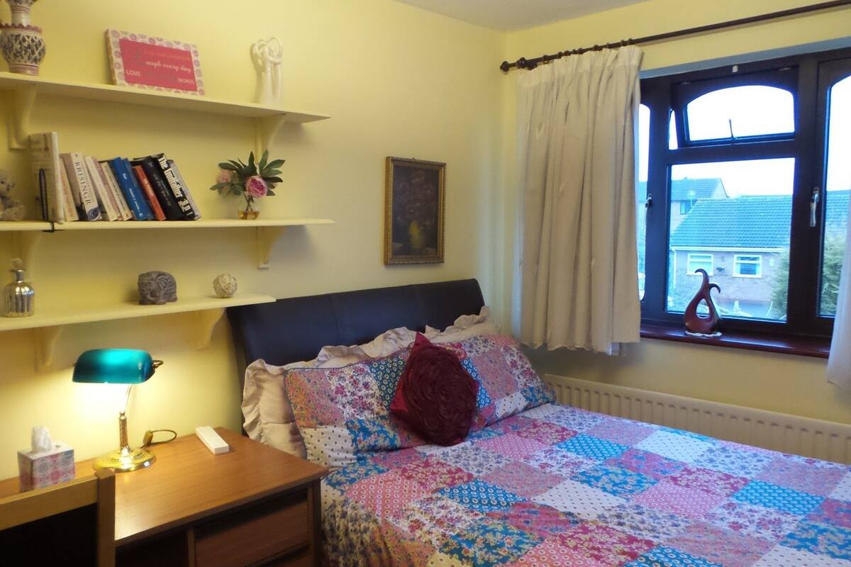 A bedroom with a bed, a desk, shelves with books and decorative items, a window with curtains, and a radiator.
