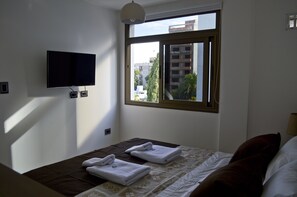 A hotel room with a bed, a TV mounted on the wall, and a window with a view of buildings and trees. - XV Apartamentos (La Plata)