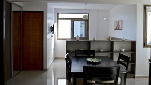 Un espace repas moderne avec une table et des chaises noires, une fenêtre avec des stores et un mur blanc orné d’un petit tableau.