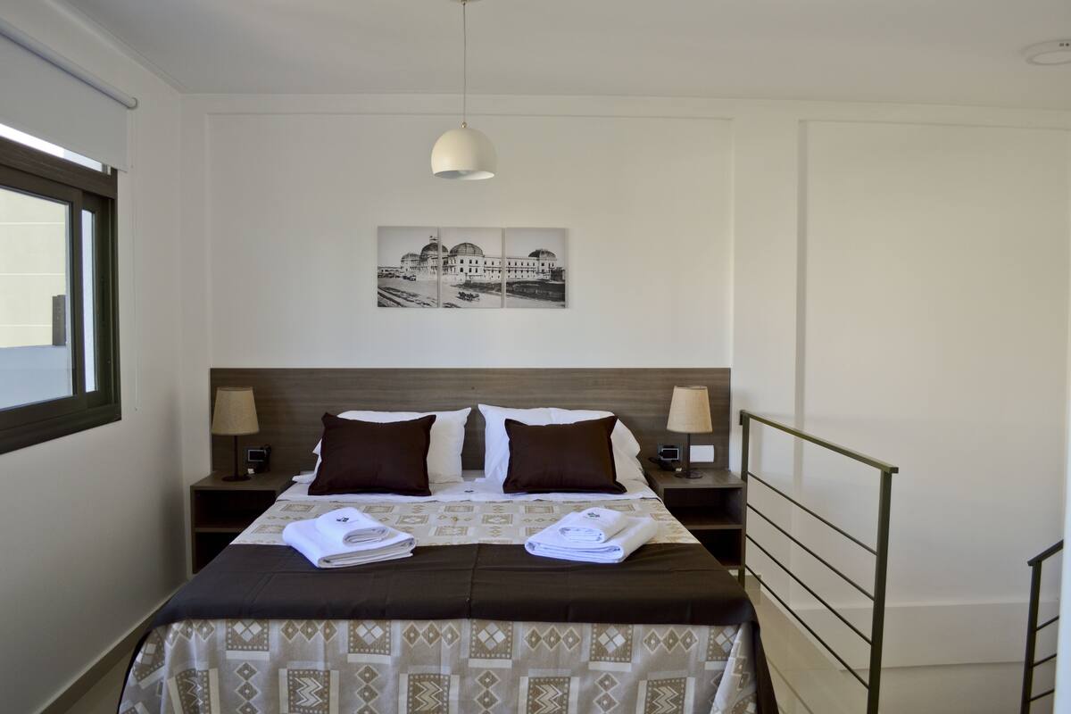 A hotel room with a bed, two nightstands, a lamp, a picture on the wall, and a window.