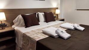 Un lit bien fait, recouvert d’une couvre-lit à motifs, avec deux oreillers blancs et deux oreillers bruns. On y trouve deux tables de chevet avec des lampes, un tableau accroché au mur et une fenêtre avec des stores.