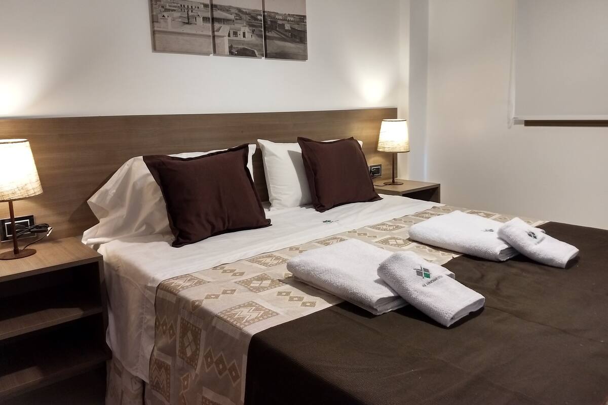 A neatly made bed with a patterned bedspread, two white pillows, and two brown pillows. There are two bedside tables with lamps, a wall-mounted picture, and a window with blinds.
