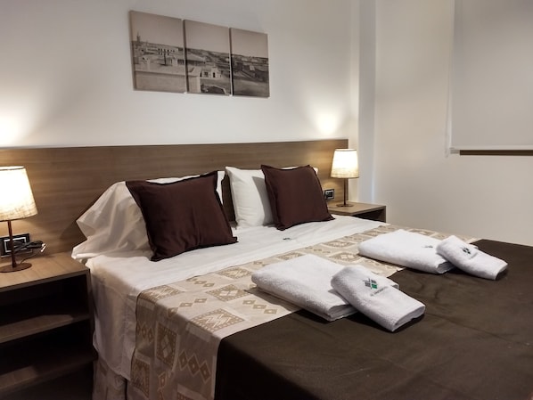 A neatly made bed with a patterned bedspread, two white pillows, and two brown pillows. There are two bedside tables with lamps, a wall-mounted picture, and a window with blinds.