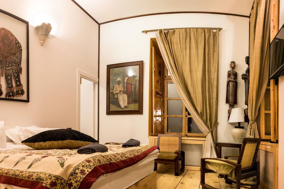 A bedroom with a bed, a chair, a desk, a lamp, and a painting of an elephant.