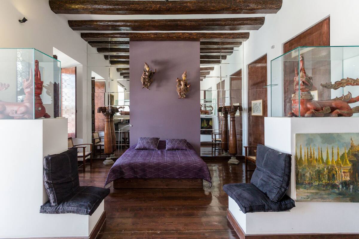 A bedroom with a bed, two chairs, a glass display case with sculptures, and a painting on the wall.