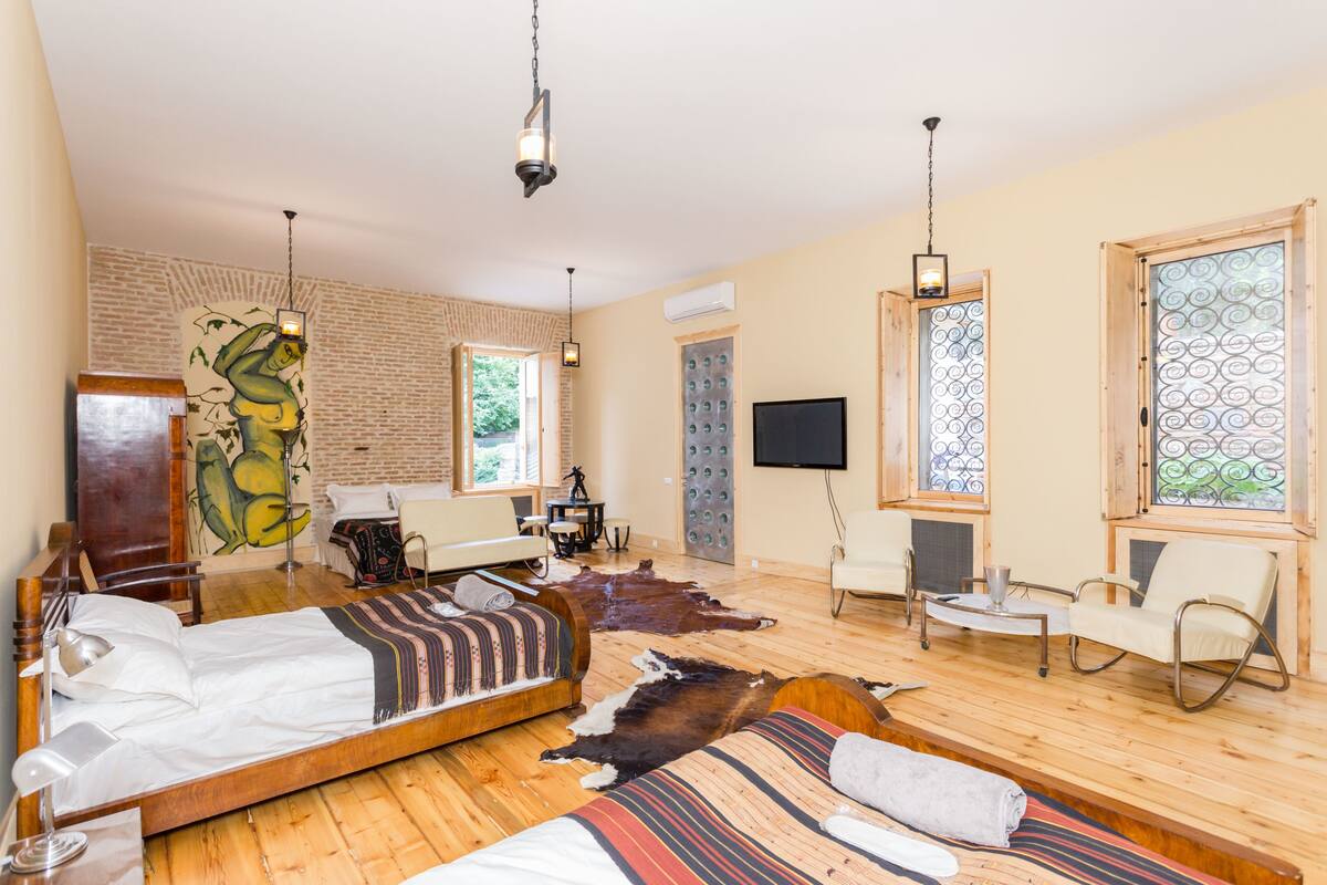A spacious room with a large bed, a desk, and a TV. There are wooden floors and a brick wall.