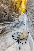 A narrow outdoor space with a glass table and wicker chair, surrounded by rocky walls and a narrow passageway.