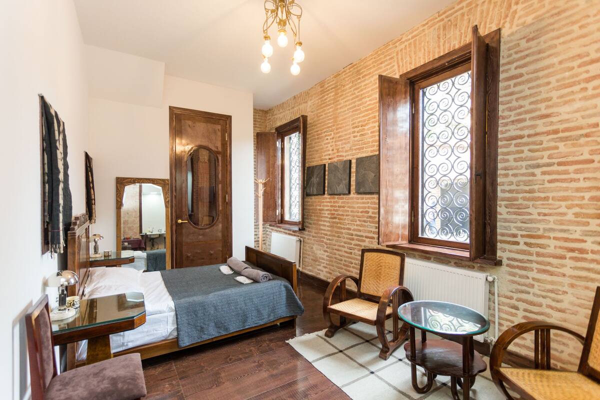 A bedroom with a wooden bed, a chair, a small table, and a window with decorative glass.