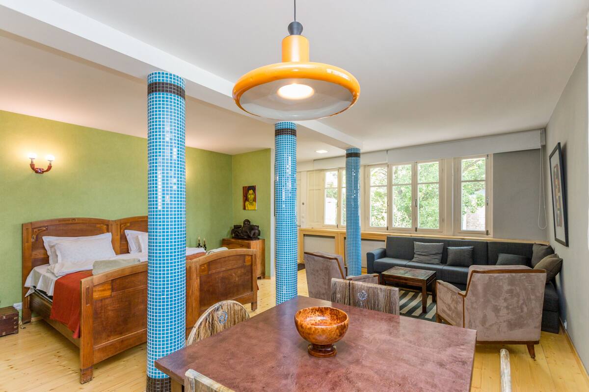 A room with a wooden bed, a dining table with a bowl, a sofa, and a large window.