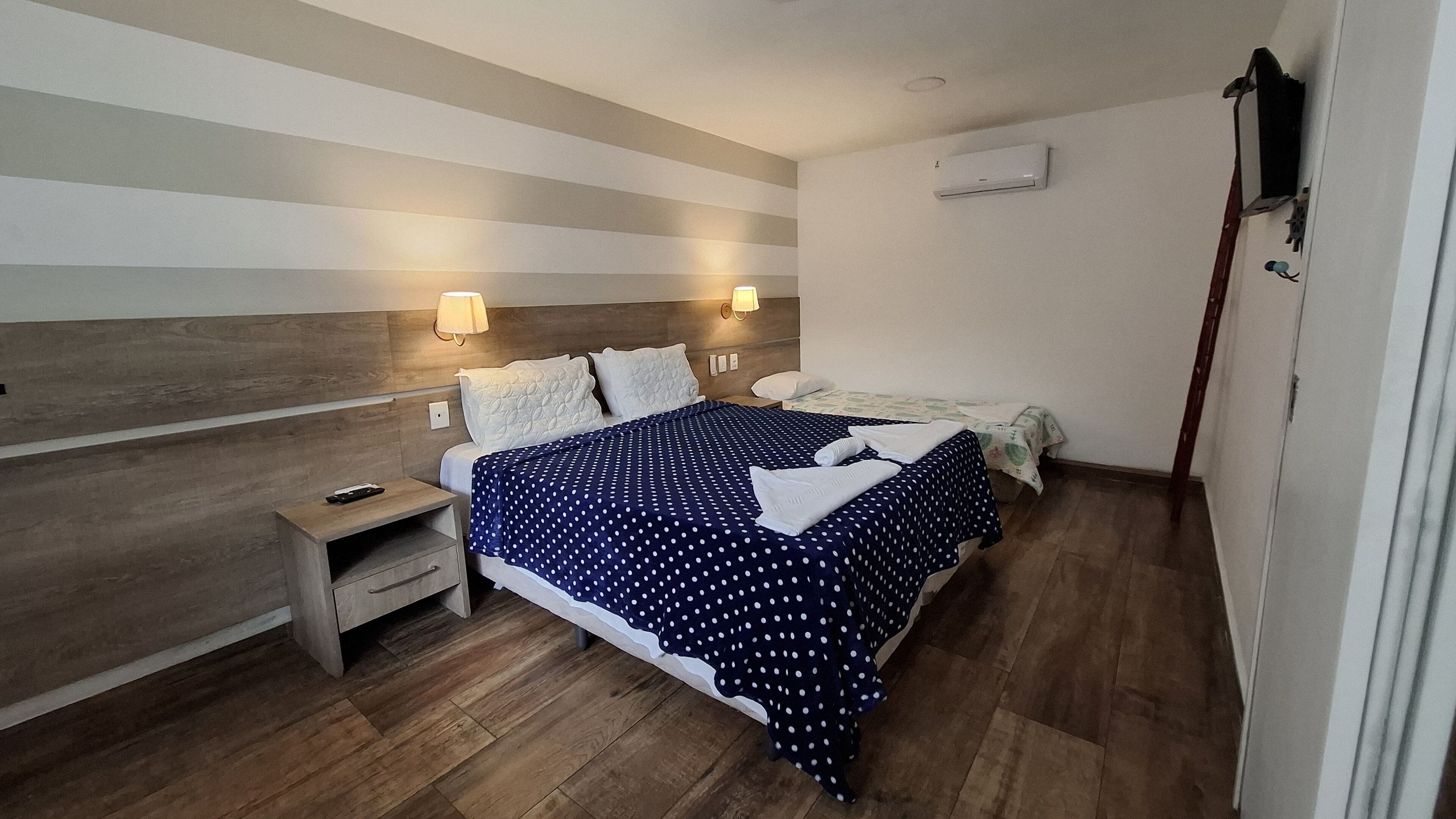A hotel room with a wooden headboard, a bed with a polka dot bedspread, two bedside lamps, a nightstand with a remote control, and an air conditioning unit on the wall.