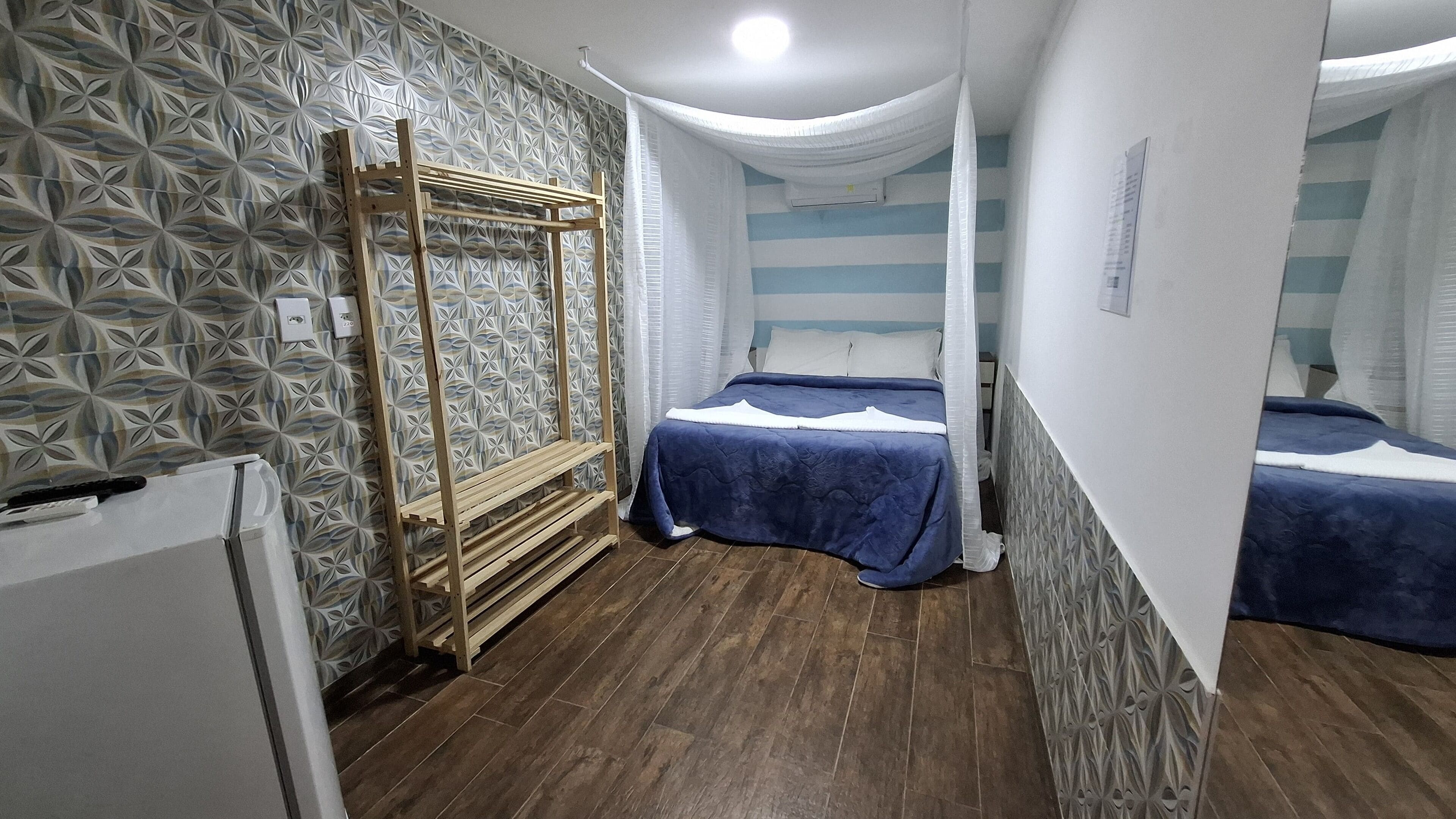 Ein Zimmer mit rautenmusterbehang, einem Bett mit blauer Bettdecke, einer hölzernen Sitzbank und einer weißen Deckenleuchte.