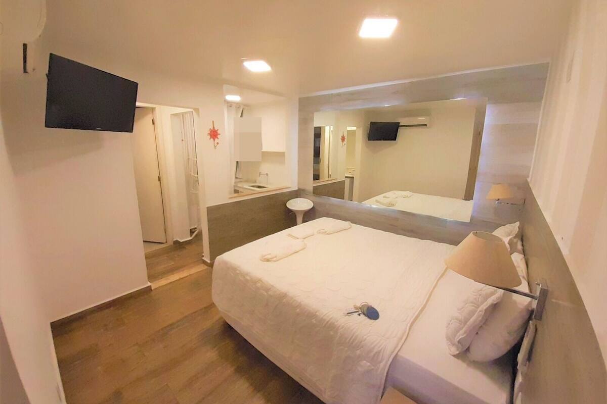 A hotel room with a bed, a bedside table, a mirror, a TV mounted on the wall, and a bathroom visible through an open door.