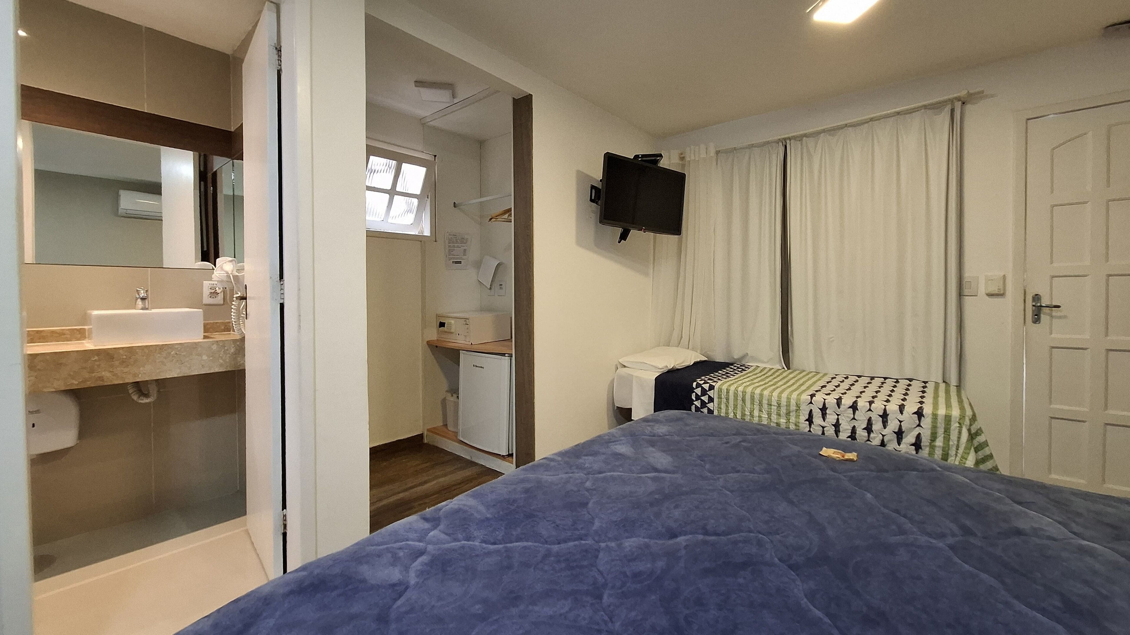 A bedroom with a bed, a bedside table, a flat-screen TV mounted on the wall, and a window with curtains.