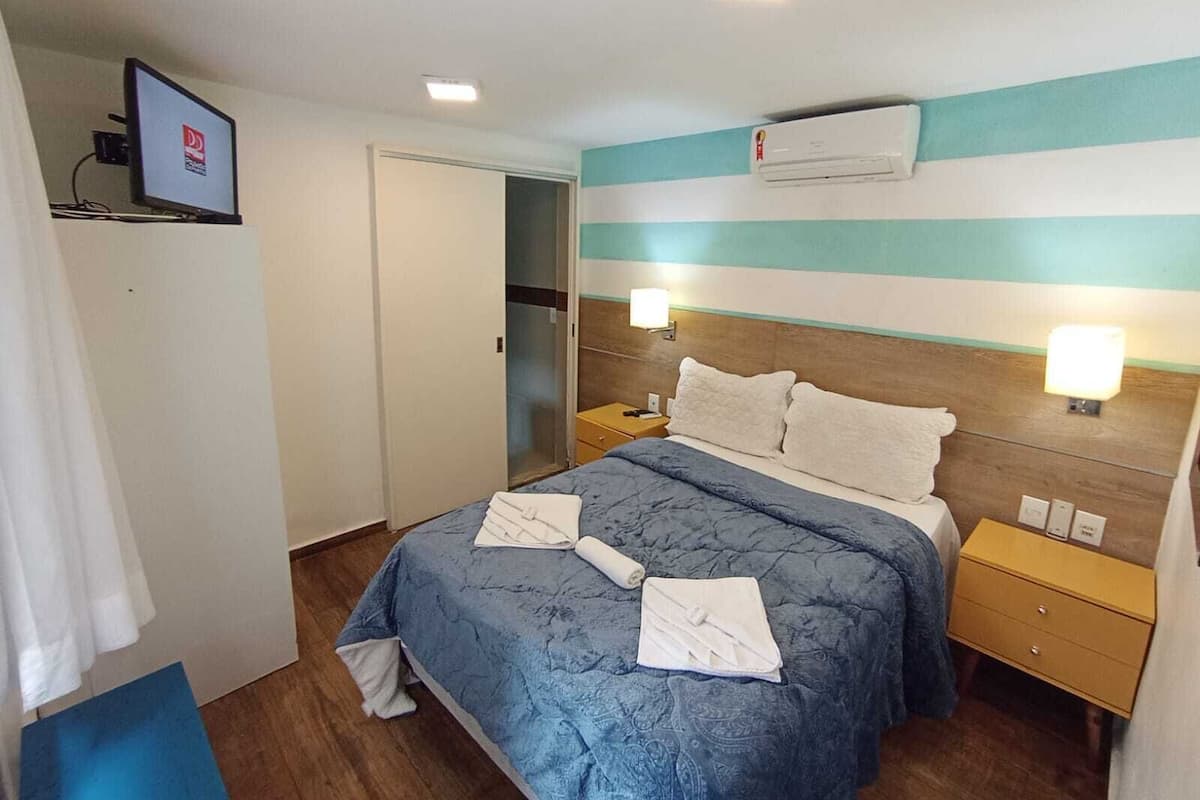 A hotel room with a bed, a TV mounted on the wall, an air conditioning unit above the bed, and a nightstand with a lamp.