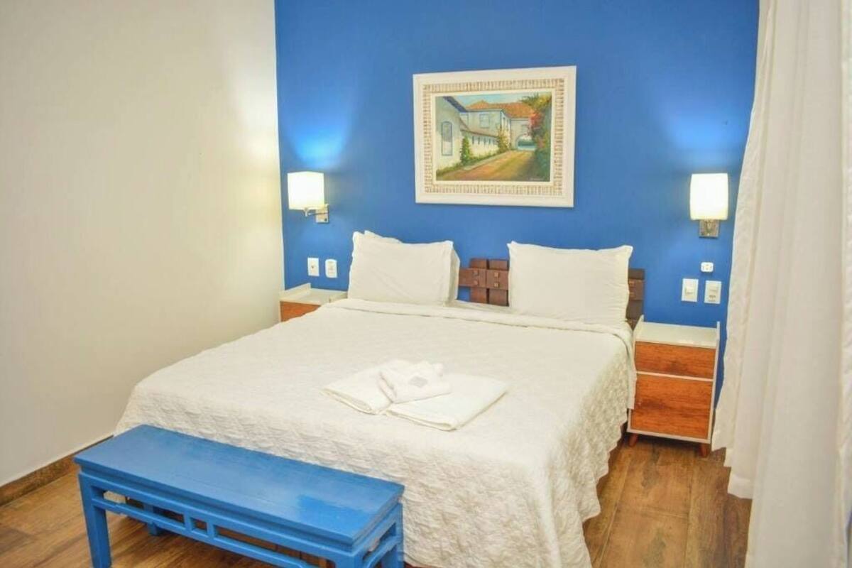 A bedroom with a large bed, two bedside tables, a blue bench, and a painting on the wall.